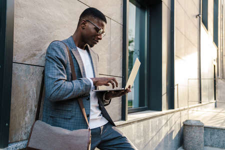 Handsome smiling smart african man wearing suit using laptop computer while standing in the streetの写真素材