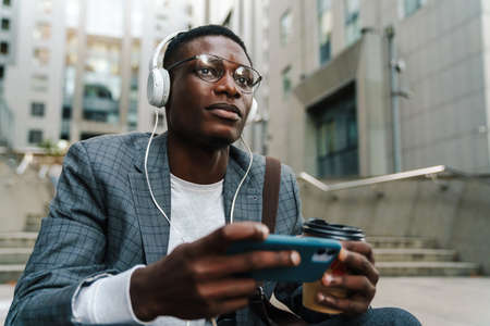 Pleased african american man in headphones using mobile phone and drinking coffee on city street