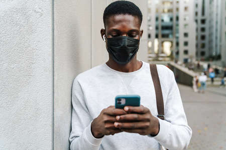Young african american man in face mask using earphones and mobile phone outdoorsの写真素材
