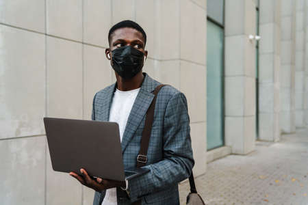 Focused african american man in face mask using laptop and earphones outdoorsの写真素材