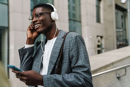 Attractive smiling young african businessman in headphones holding mobile phone while walking in the streetの写真素材