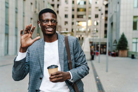 Happy african american man in earphones waving hand while drinking coffee on city streetの写真素材