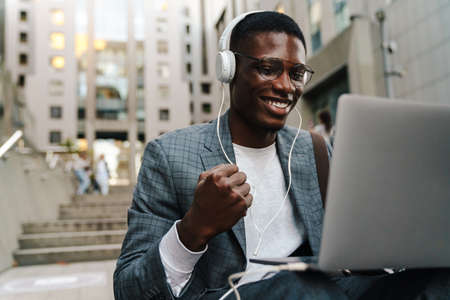 Cheerful african american man in headphones using laptop while making winner gesture outdoorsの写真素材