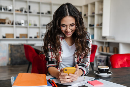 Joyful nice woman using cellphone and working with papers while drinking coffee in cafe