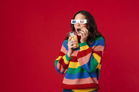 Shocked beautiful girl eating popcorn and watching movie isolated over red backgroundの写真素材
