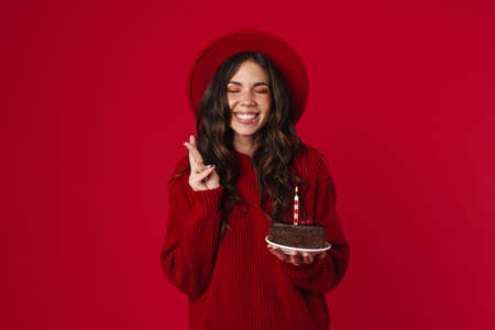 Cheerful beautiful girl posing with birthday cake and fingers crossed isolated over red background
