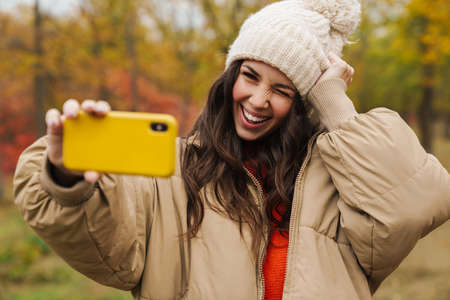 Brunette happy woman winking and smiling while taking selfie on cellphone in autumn forestの写真素材