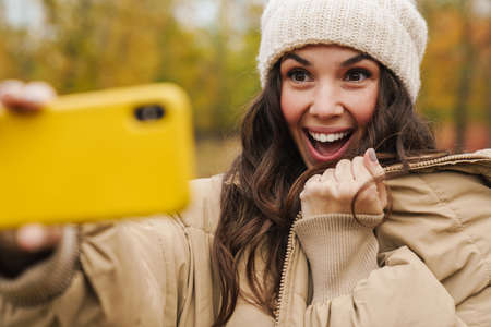 Brunette excited woman in knit hat taking selfie on cellphone while strolling in autumn forest