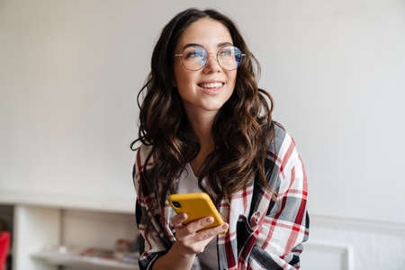 Happy nice brunette woman smiling and using mobile phone indoorsの写真素材