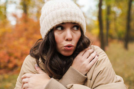 Beautiful unhappy woman in knit hat trembling while strolling in autumn forest