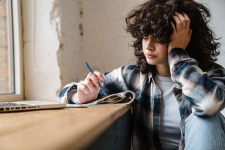Pensive young woman taking notes while sitting at the window indoors