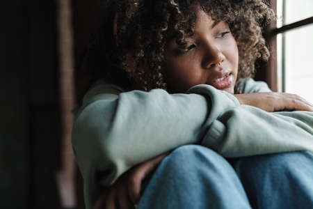 Focused african american woman looking at window while sitting on windowsill indoors
