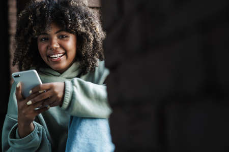 Joyful african american woman using mobile phone while sitting on windowsill indoors