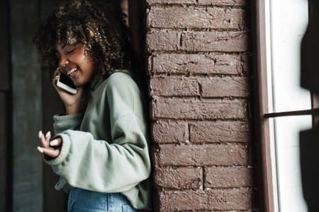 Joyful african american woman laughing and talking on mobile phone while leaning on wall indoorsの写真素材