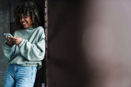 Joyful african american woman smiling and using mobile phone while leaning on wall indoors