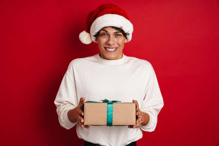 Handsome smiling guy in Santa Claus hat posing with Christmas gift isolated over red background