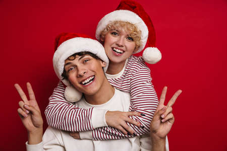 Beautiful happy couple in Santa Claus hats showing peace sign isolated over red backgroundの写真素材