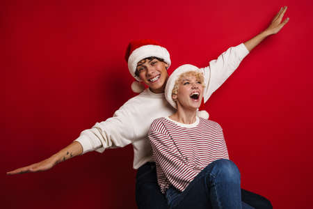Beautiful happy couple in Santa Claus hats making fun at camera isolated over red background