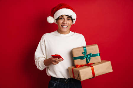 Handsome smiling guy posing with smarphone and Christmas gifts isolated over red background