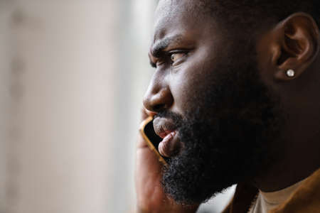 Displeased african american man posing and talking on mobile phone indoorsの写真素材