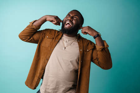 Displeased african american man screaming and plugging his ears isolated over blue background