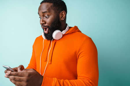 Surprised african american man with headphones using cellphone isolated over blue background