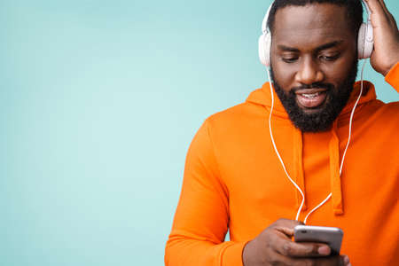 Cheerful african american man using cellphone and headphones isolated over blue background