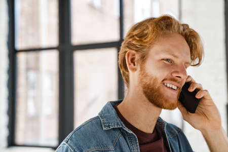 Happy handsome ginger guy smiling and talking on mobile phone indoorsの写真素材