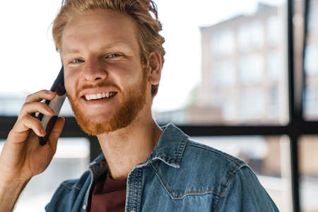 Happy handsome ginger guy smiling and talking on mobile phone indoors