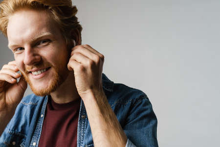 Joyful redhead guy in wireless earphones smiling at camera isolated over grey background