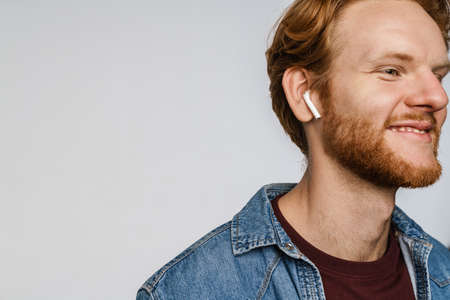 Joyful redhead guy in wireless earphones smiling at camera isolated over white background