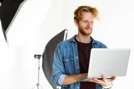 Pleased handsome ginger guy smiling while working with laptop in studio indoorsの写真素材