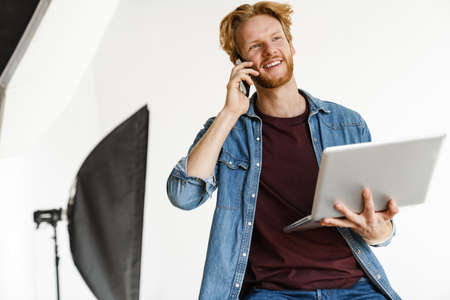 Smiling male photographer using laptop computer while sitting at the studio, talking on mobile phone
