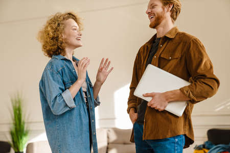 Attractive smiling young couple talking while standing indoors, carrying laptop computerの写真素材