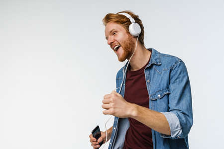 Happy handsome redhead guy in headphones using cellphone and dancing isolated over white background