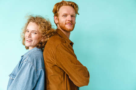 Young happy redhead couple smiling while posing back to back isolated over blue background