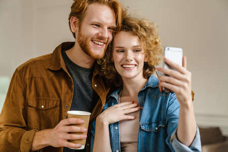 Young happy redhead couple drinking coffee and taking selfie on mobile phone indoorsの写真素材
