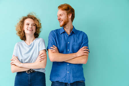 Young happy redhead couple laughing while posing with arms crossed isolated over blue background