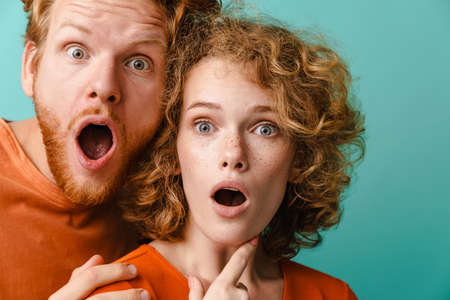 Young shocked redhead couple expressing surprise at camera isolated over blue background