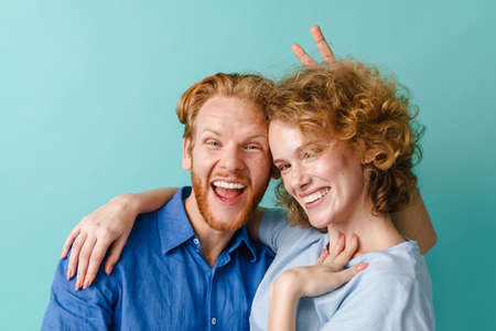 Young cheerful redhead couple making fun and horns with fingers isolated over blue backgroundの写真素材