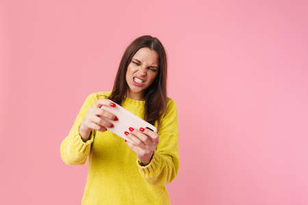 Displeased brunette girl playing online game on mobile phone isolated over pink background