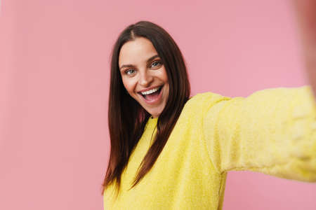 Happy beautiful girl smiling while taking selfie photo isolated over pink background