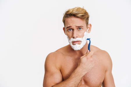 Unhappy caucasian shirtless man shaving with foam and razor isolated over white backgroundの写真素材