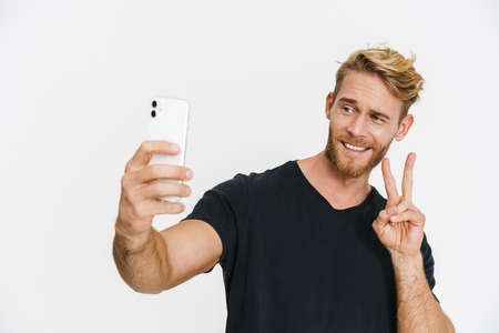 Smiling blonde man gesturing peace sign while taking selfie on cellphone isolated over white...