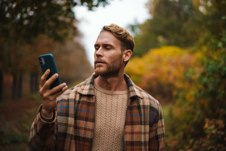 Handsome unshaven man using mobile phone while strolling in autumn forestの写真素材