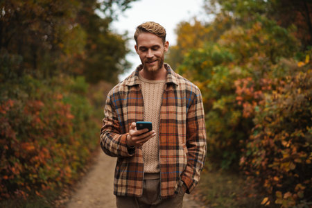 Joyful unshaven man using mobile phone while strolling in autumn forestの写真素材