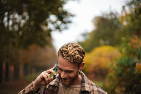 Joyful unshaven man talking on mobile phone while strolling in autumn forestの写真素材