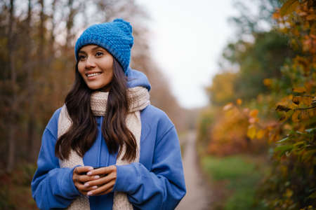 Nice brunette happy woman in knit hat drinking tea while strolling in autumn forest