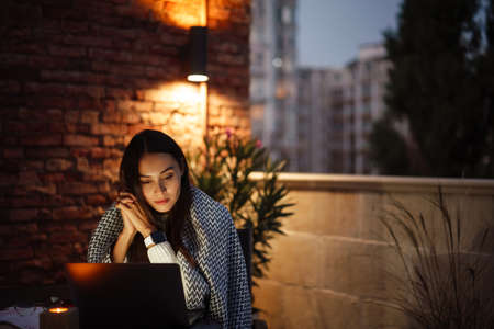 Attractive young asian woman covered in blanket using laptop computer while sitting at the balcony...