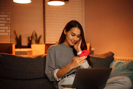 Smiling beautiful woman using cellphone while working with laptop on sofa at homeの写真素材